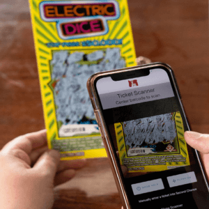 in the background, a hand holds an Electric Dice Scratch-it. In the foreground, the other hand holds a smartphone and is using the lottery app to scan the ticket's second chance code